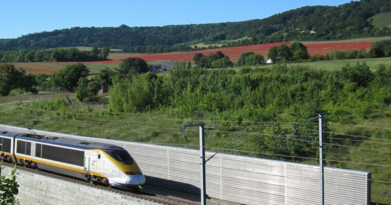 A high-speed train moves along an elevated track through a lush countryside. The scene is framed by greenery, rolling hills, and fields with patches of red under a clear blue sky.