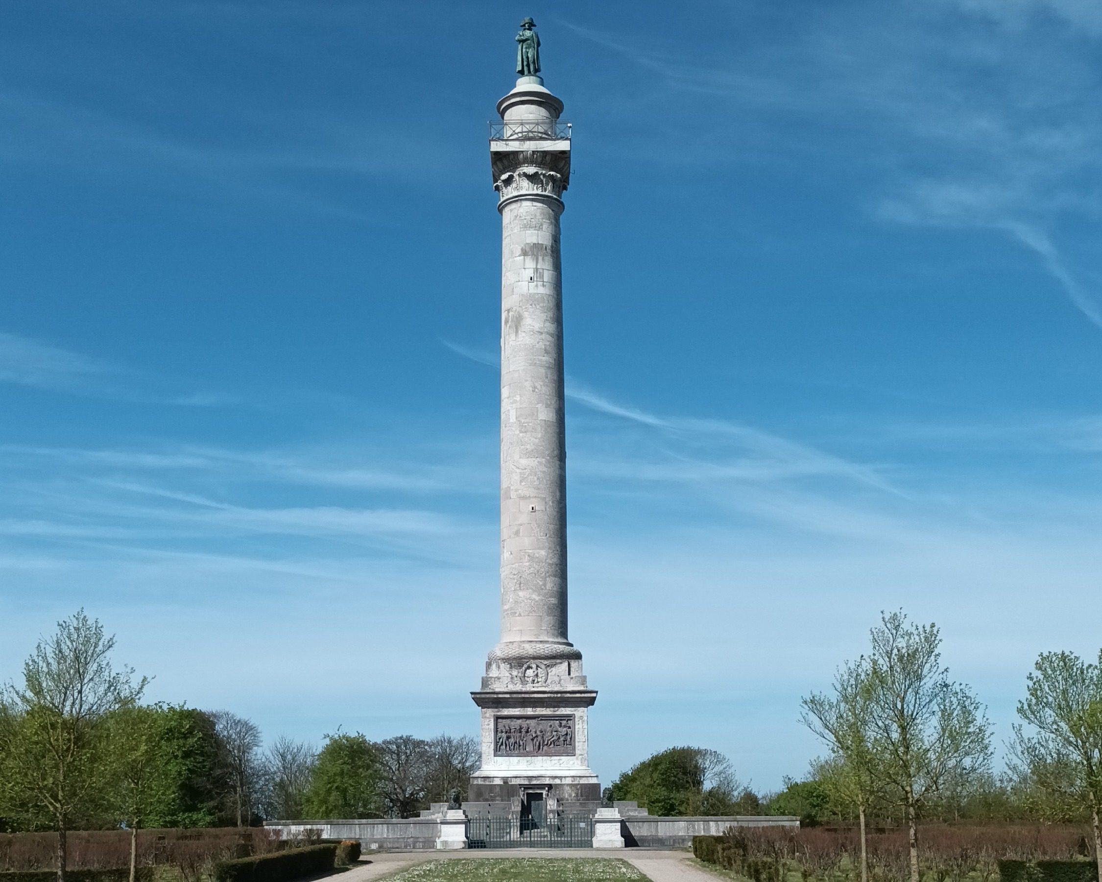A tall stone column with a statue on top, set against a clear blue sky. The statue depicts a standing figure, possibly a historical or cultural icon. The column features decorative carvings near its upper section.