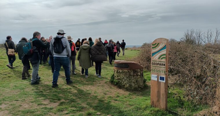 A group of people walking through a grassy landscape towards a stone structure with an informational sign reading "Cross-Channel Geopark Transfrontier Geosite." The sign features a European Union flag and logo. The sky is overcast, and trees and bushes frame the background. This scene highlights exploration within a geopark, emphasizing geological significance and natural beauty.