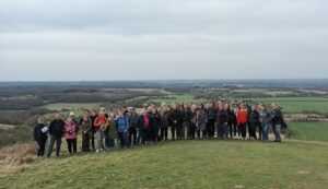 A large group of people gathered on a grassy hilltop, enjoying a scenic view of the countryside. The landscape features fields, forests, and distant hills under a cloudy sky, suggesting an outdoor excursion or group hike.