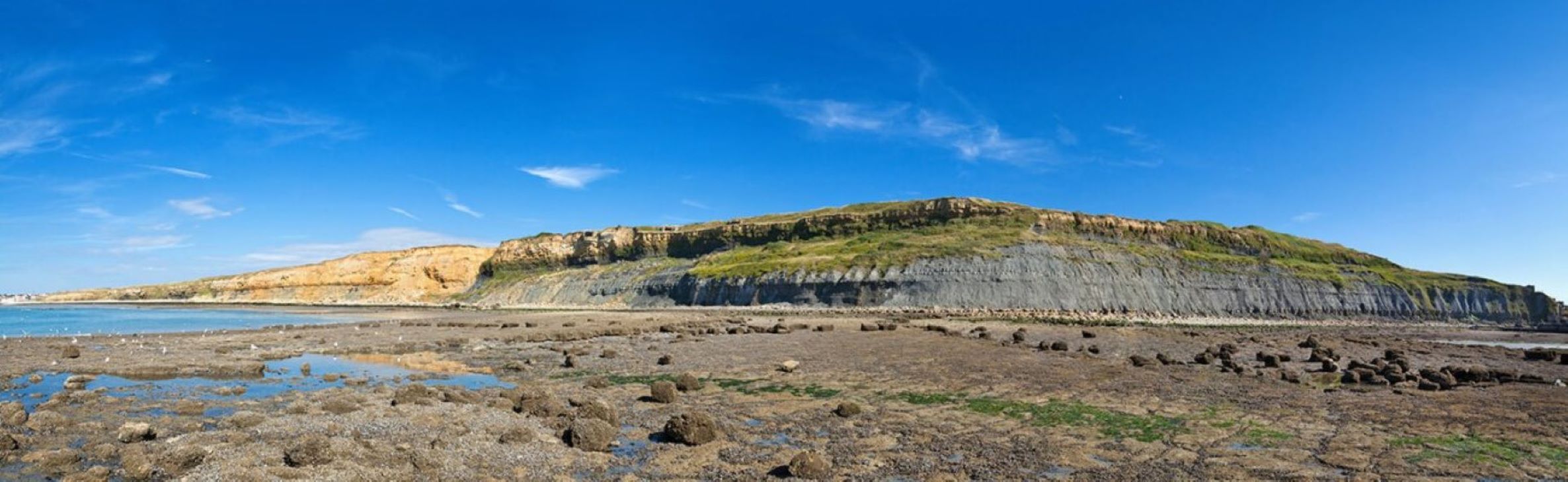 A coastal landscape featuring a sandy beach with a shallow pool of water. The backdrop includes a rugged cliff with visible layers of sediment and rock, showcasing geological formations. The sky is mostly clear with a few wispy clouds.