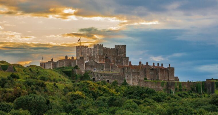 Dover Castle, a large medieval fortress with stone walls and towers, sits atop a green hill surrounded by dense vegetation under a dramatic sky with clouds and patches of golden sunlight.